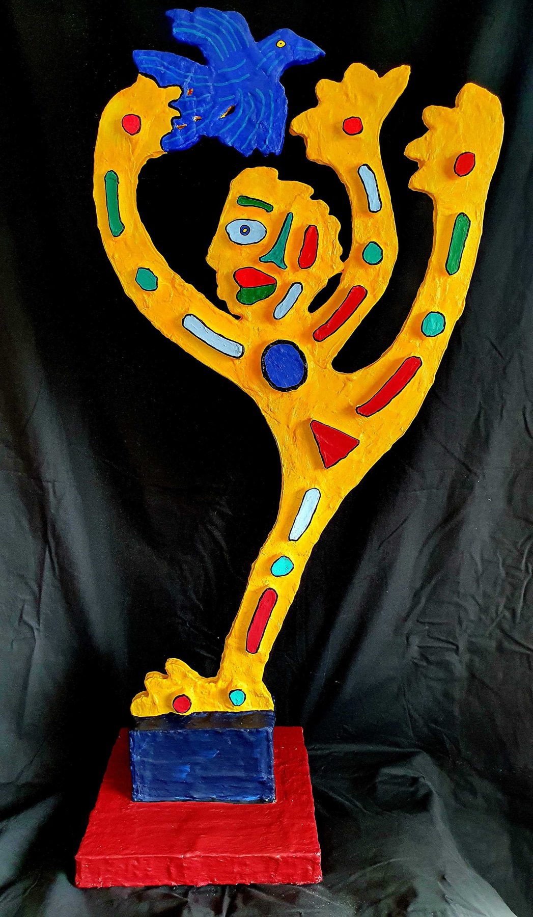 L'envol : sculpture-totem jaune, bras levés, oiseau bleu
